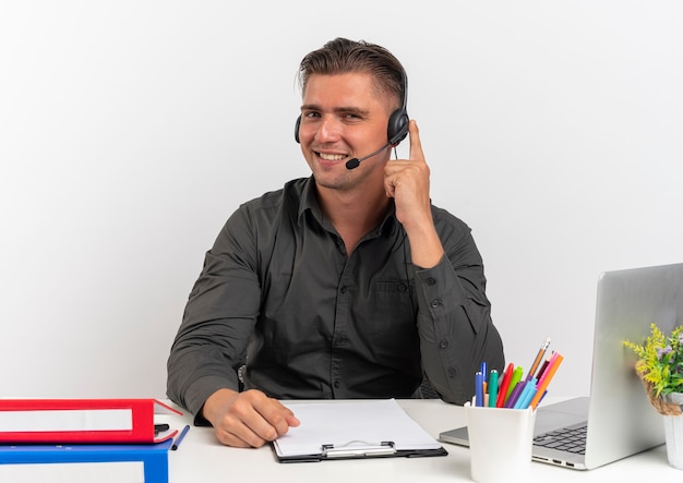 Young smiling blonde office worker man sur les écouteurs est assis au bureau avec des outils de bureau à l'aide d'un ordinateur portable met le doigt sur un casque isolé sur fond blanc avec copie