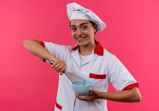 Young smiling caucasian cook girl en uniforme de chef détient bol et fouet isolé sur mur rose avec copie espace