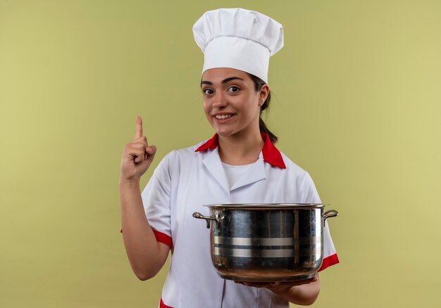 Young smiling caucasian cook girl en uniforme de chef détient pot et pointe isolé sur mur vert avec espace copie