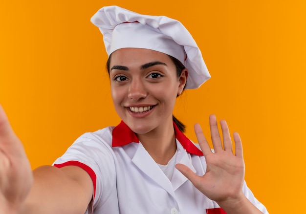 Young smiling caucasian cook girl en uniforme de chef montre la main isolée sur un mur orange