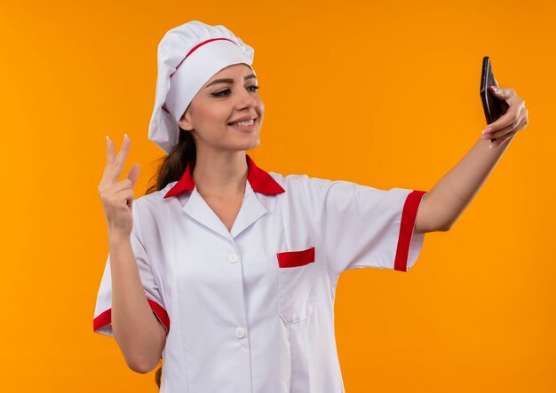 Young smiling caucasian cook girl en uniforme de chef regarde le téléphone et les gestes signe de la main de la victoire isolé sur le mur orange avec copie espace