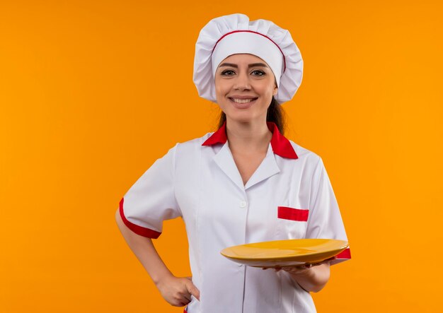 Young smiling caucasian cook girl en uniforme de chef tient la plaque et met la main sur la taille isolé sur un mur orange avec copie espace