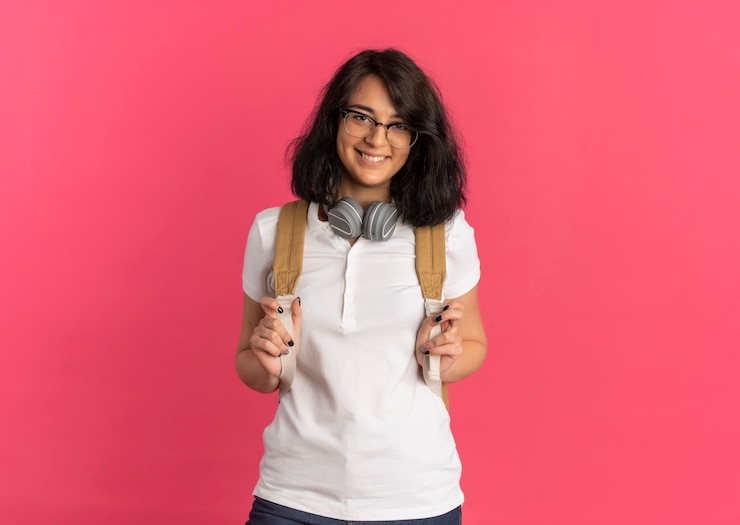 Photo gratuit young smiling jolie écolière caucasienne portant des lunettes casque sur le cou et le sac à dos regarde la caméra sur rose avec copie espace