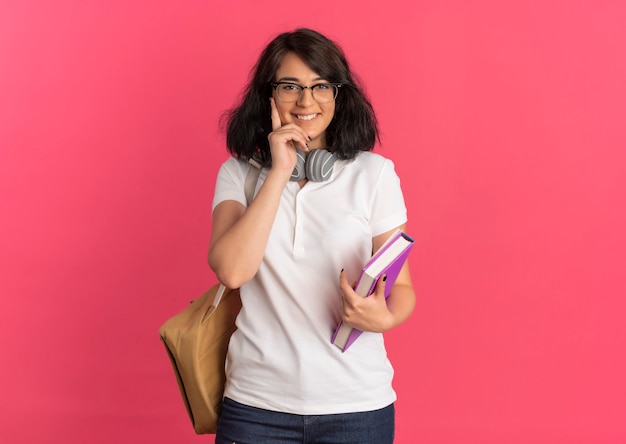 Young smiling jolie écolière caucasienne portant des lunettes et sac à dos met la main sur le visage tenant des livres sur rose avec copie espace