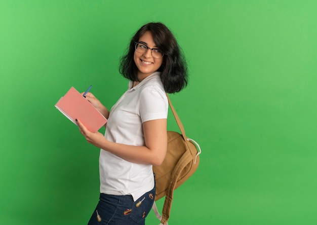 Young smiling jolie écolière caucasienne portant des lunettes et sac à dos se dresse sur le côté tenant un stylo et un cahier sur vert avec copie espace