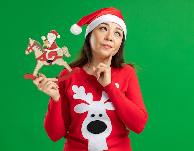 Young woman wearing christmas santa hat et pull rouge tenant le jouet de Noël à la perplexité debout sur fond vert