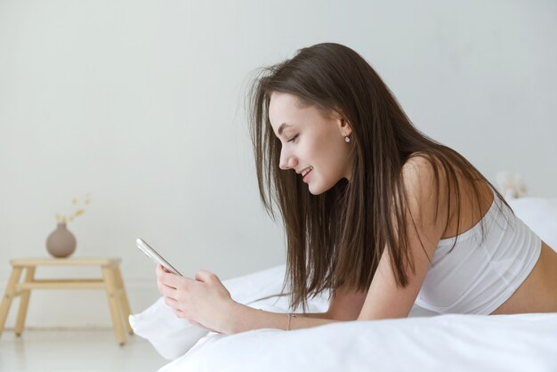 1 jeune femme brune blanche en haut blanc allongé sur le lit avec téléphone, vue latérale