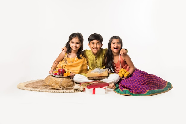 3 enfants indiens ou frères et sœurs en tenue traditionnelle assis sur un canapé ou un fond blanc, tenant des cadeaux et des bonbons ou du laddu