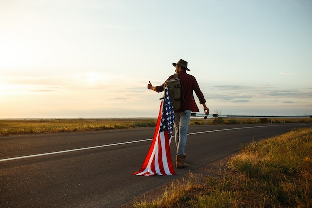 4 juillet. Le quatre juillet. Américain avec le drapeau national. Drapeau américain. Le jour de l'indépendance. Fête patriotique. L'homme porte un chapeau, un sac à dos, une chemise et un jean.