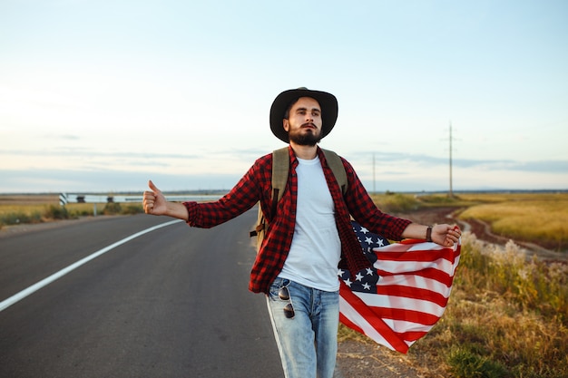 4 juillet. Le quatre juillet. Américain avec le drapeau national. Drapeau américain. Le jour de l'indépendance. Fête patriotique. L'homme porte un chapeau, un sac à dos, une chemise et un jean.