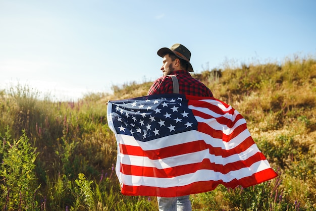 4 juillet. Le quatre juillet. Américain avec le drapeau national. Drapeau américain. Le jour de l'indépendance. Fête patriotique. L'homme porte un chapeau, un sac à dos, une chemise et un jean.