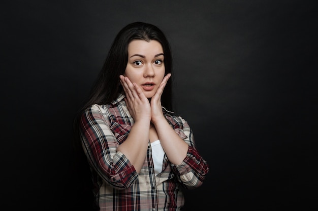 Accablé jeune fille mignonne démontrant des émotions confuses et levant la main à la tête en se tenant debout isolé dans le mur noir