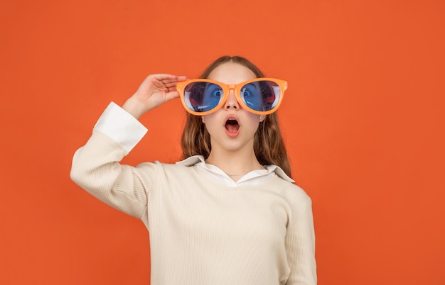 Accessoire de mode. devenir fou. bonheur d'enfance. look de fête chic. joyeux enfant drôle dans des lunettes glamour sur fond orange. adolescente géniale s'amusant. enfant surpris dans des verres de fête.