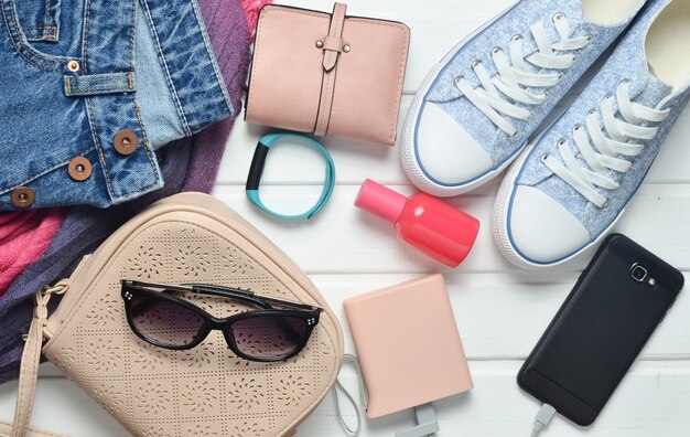 Accessoires, chaussures, vêtements et gadgets modernes pour femmes sur un fond en bois blanc. Jeans, sac, baskets, smartphone, bracelet intelligent, banque d'alimentation, cosmétiques, lunettes de soleil, écharpe. Vue de dessus.