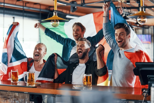 Acclamations de jeunes hommes couverts de drapeaux internationaux dégustant de la bière tout en regardant un match de sport au pub