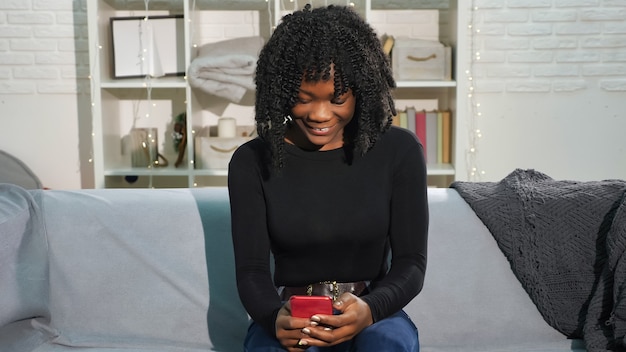 Accro aux médias sociaux, une jolie fille afro-américaine regarde un smartphone rouge à la maison