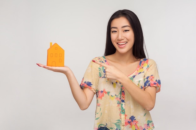 Achat de propriété et assurance. Belle fille asiatique heureuse avec des cheveux bruns en chemisier d'été pointant vers la maison de papier sur la paume et souriant avec contentement. studio d'intérieur tourné isolé sur fond gris