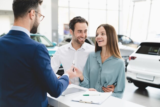 Photo achat d'une voiture neuve assistant de magasin masculin félicitant un jeune couple d'acheteurs d'automobiles en donnant les clés de la voiture au magasin du concessionnaire