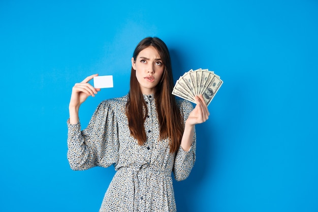 Achats. Fille réfléchie regardant et mordant la lèvre, en comptant à l'esprit ou en pensant, tenant une carte de crédit en plastique avec des billets d'un dollar, debout sur le bleu.