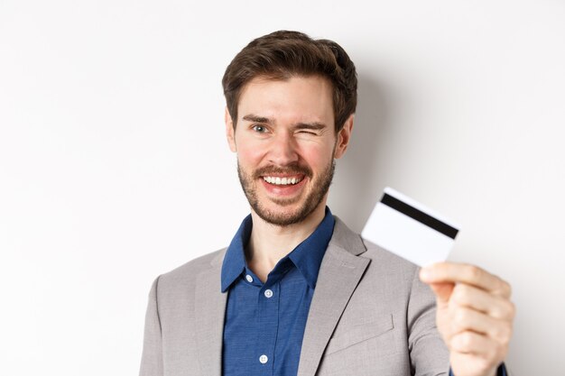 Achats. Gai barbu en costume clignotant et souriant, montrant une carte de crédit en plastique à la caméra, fond blanc.