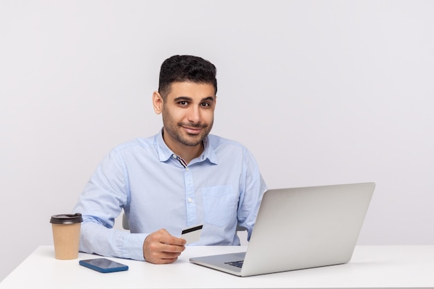 Achats sur Internet commerce électronique Heureux homme d'affaires élégant assis sur le lieu de travail avec un ordinateur portable détenant une carte de crédit et souriant à la caméra faisant un achat en ligne tourné en studio isolé sur fond blanc