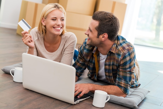 Les achats en ligne facilitent la vie. Jeune couple souriant allongé sur le sol de leur nouvel appartement et faisant des achats sur Internet tandis que des boîtes en carton sont posées en arrière-plan