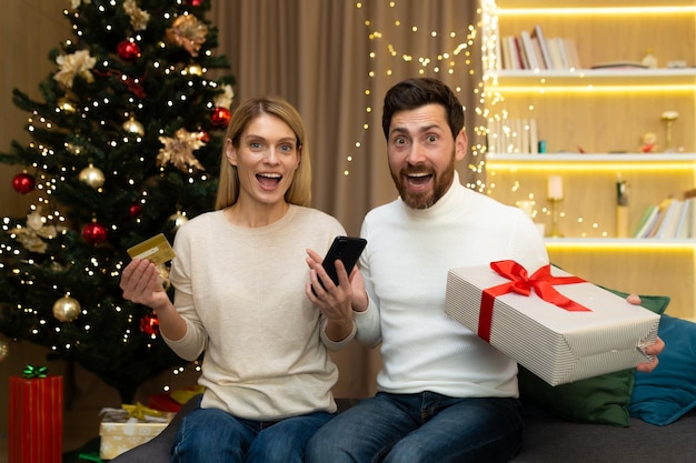 Achats en ligne heureux jeune couple à la maison près de l'arbre de noël tenant une carte de crédit téléphone et cadeau nous