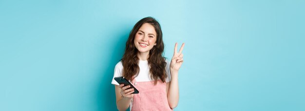 Achats en ligne Jolie et joyeuse jeune femme souriante tenant un smartphone et montrant la paix vsign à la caméra debout sur fond bleu
