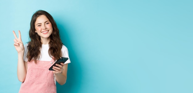 Achats en ligne Jolie et joyeuse jeune femme souriante tenant un smartphone et montrant la paix vsign à la caméra debout sur fond bleu
