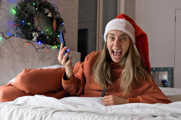 Photo achats en ligne de noël : une femme heureuse dans un bonnet de noel rouge avec smartphone. cadeaux via les boutiques en ligne.