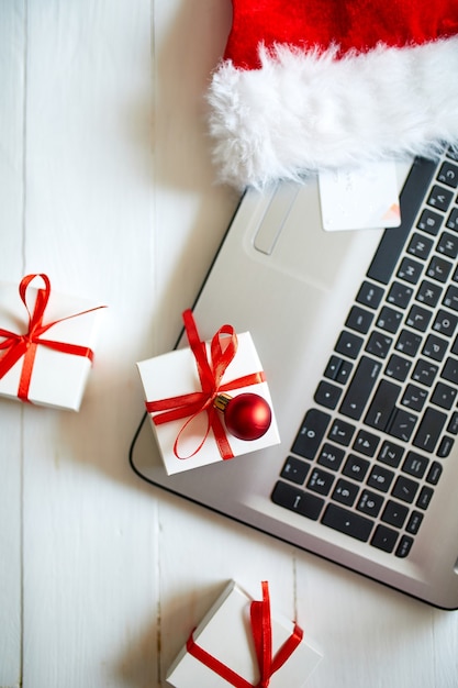 Achats en ligne de Noël, ordinateur portable, carte de crédit, bonnet de Noel et boîte-cadeau sur table en bois blanc, vue du dessus