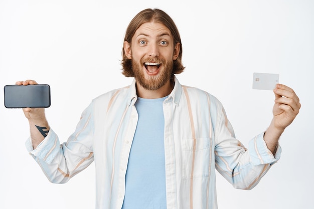 Achats en ligne, technologie. Homme souriant excité montrant l'écran horizontal du téléphone portable et la carte de réduction de crédit, haletant étonné, recommandant l'application, fond blanc.