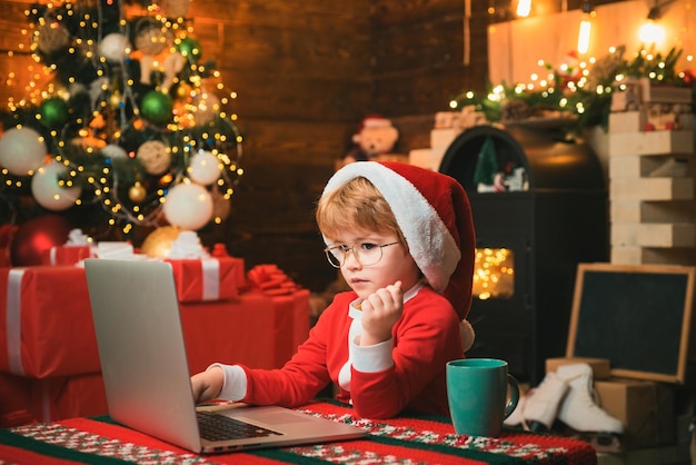 Achats de Noël en ligne pour les enfants Enfant de Noël Heureux petit garçon souriant portant des vêtements de Père Noël et écrivant une lettre sur son ordinateur portable