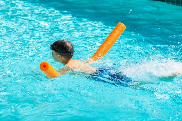 Activité estivale en plein air Concept de santé et de vacances amusantes Heureux garçon souriant de huit ans dans des lunettes de natation nager dans la piscine avec des nouilles dans une chaude journée d'été