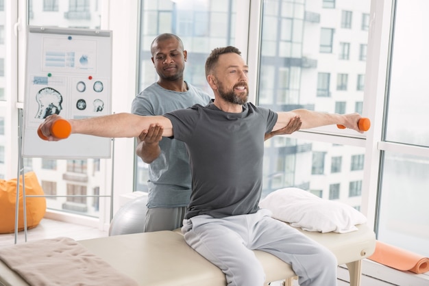 Activité de rééducation. Enthousiaste bel homme souriant tout en exerçant avec des haltères