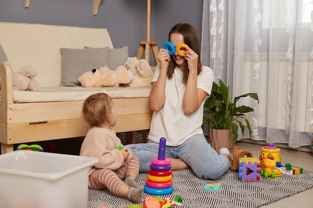 Activités avec des enfants à la maison développement de la petite enfance Jeune adulte femme assise sur le sol dans le salon jouant avec son bébé avec une pyramide couvrant ses yeux avec des anneaux s'amusant