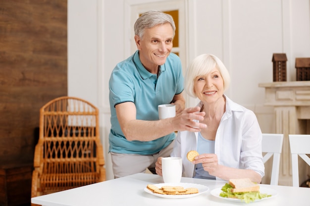 Admirable homme attentif inspiré pointant vers la fenêtre alors que lui et sa femme apprécient un café frais tout en prenant le petit déjeuner