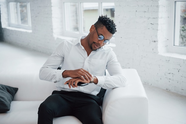 Admire la nouvelle montre-bracelet. Guy afro américain élégant en vêtements classiques et lunettes est assis sur le lit