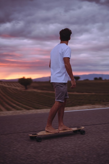 Adolescent actif faisant du skateboard sur la route et admirant le paysage pittoresque au coucher du soleil contre un ciel nuageux. Garçon sportif faisant de la planche à roulettes tout en regardant une vue idyllique sur le terrain pendant le coucher du soleil
