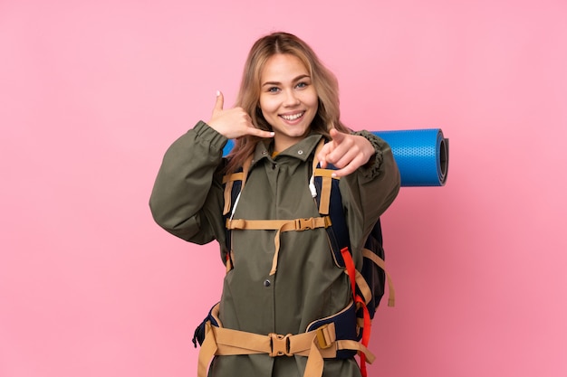 Adolescent alpiniste fille avec un gros sac à dos isolé sur le mur rose faisant un geste de téléphone et pointant vers l'avant