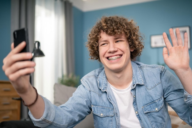 Un adolescent attrayant et souriant s'assoit sur un canapé à la maison et discute avec un ami par smartphone