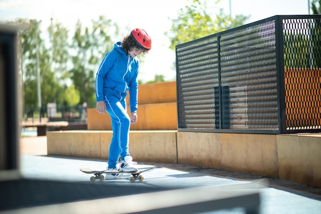 Adolescent aux cheveux bouclés souriant et satisfait dans un casque de protection mettant son pied droit sur la planche pendant l'entraînement de planche à roulettes