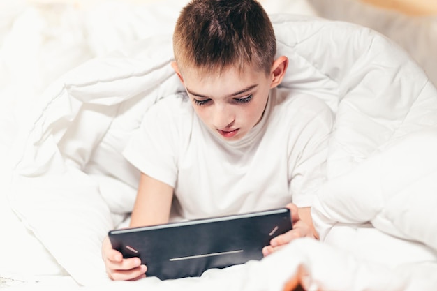 Un adolescent de bébé couché dans son lit sous une couverture blanche apprend à jouer sur une tablette tactile, un ordinateur ou un téléphone