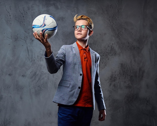 Un adolescent blond vêtu d'un jean et d'une veste grise tient un ballon de football sur fond gris.