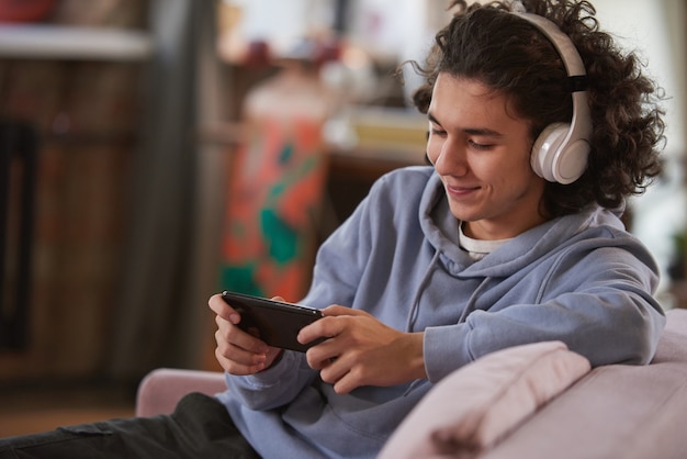 Adolescent en casque sans fil parlant en ligne à l'aide de son téléphone intelligent assis sur un canapé