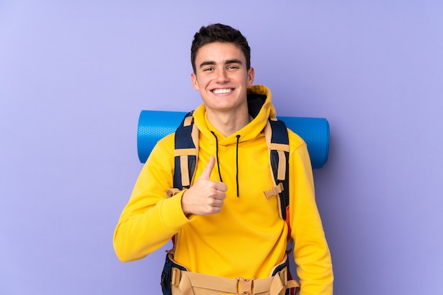 Adolescent caucasien alpiniste homme avec un gros sac à dos sur le mur violet donnant un coup de pouce geste