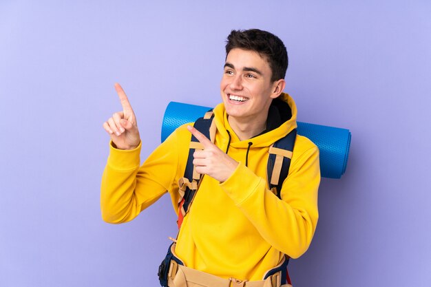 Adolescent caucasien alpiniste homme avec un gros sac à dos sur le mur violet pointant avec l'index une excellente idée