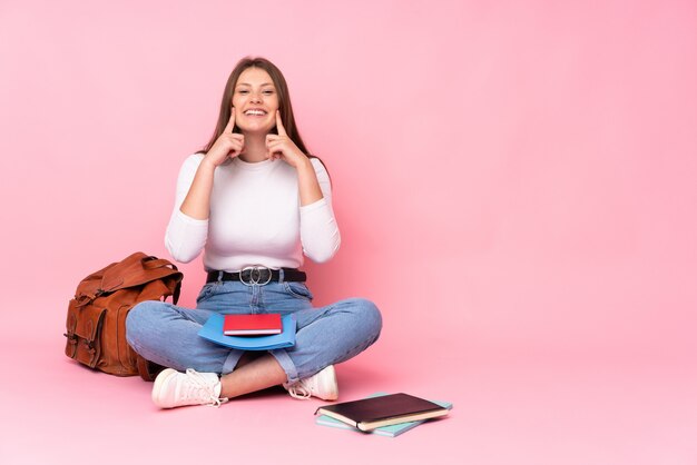 Adolescent caucasien étudiant fille assise sur le sol isolé sur mur rose souriant avec une expression heureuse et agréable