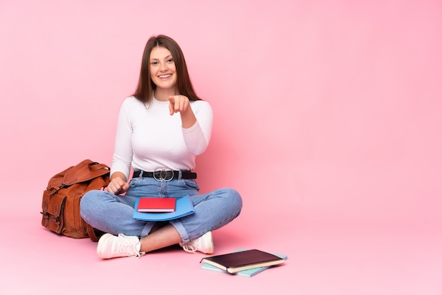 Adolescent caucasien étudiant fille assise sur le sol isolé sur le mur rose surpris et pointant vers l'avant