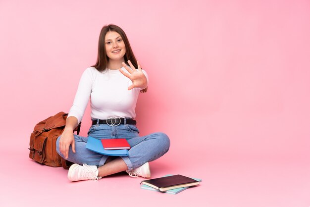 Adolescent caucasien étudiant fille assise sur le sol isolé sur rose en comptant cinq avec les doigts